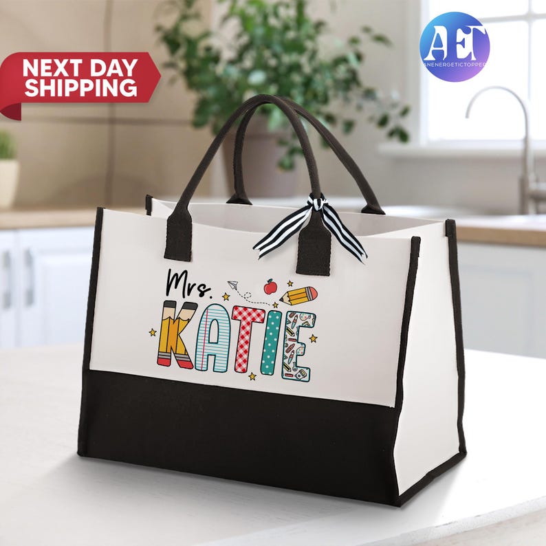 May include: White tote bag with black handles and a black bottom. The bag has a design of the words "Mrs. Katie" in colorful letters with school supplies and a red apple.
