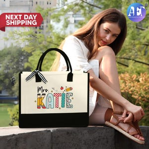 May include: A white canvas tote bag with black handles and a black bottom. The bag has a colorful design with the text "Mrs. Katie" and school-themed illustrations.