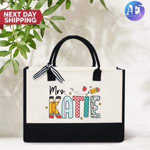 May include: A white canvas tote bag with black handles and a black bottom. The bag has a colorful design with the text "Mrs. Katie" and school-themed illustrations.