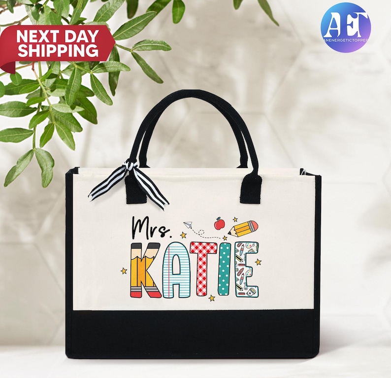May include: A white canvas tote bag with black handles and a black bottom. The bag has a colorful design with the text "Mrs. Katie" and school-themed illustrations.