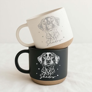 May include: Two ceramic mugs, one cream and one black, stacked. Each mug features a detailed dog portrait and the name "Shadow" in script. The mugs have a brown base and a curved handle. The cream mug has a white handle.