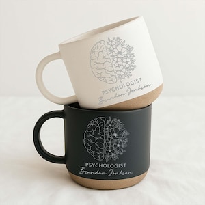 May include: Two ceramic mugs stacked on top of each other. The top mug is off-white, and the bottom mug is black. Each mug features a design of a brain and flowers, with the text "PSYCHOLOGIST Brandon Jonhson".