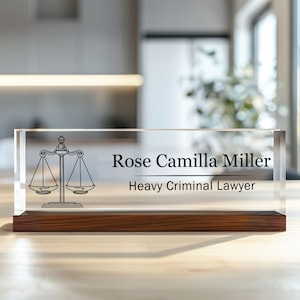 May include: A clear acrylic desk nameplate with a wooden base. The nameplate features a scale of justice design and the text "Rose Camilla Miller, Heavy Criminal Lawyer".