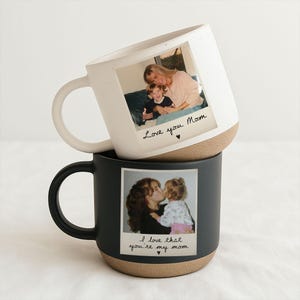 May include: Two ceramic mugs, one white and one black, stacked. Each mug features a photograph of a mother and child. The white mug says "Love you Mom" and the black mug says "I love that you're my mom."