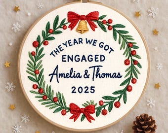 May include: Embroidered hoop art with the text "The Year We Got Engaged Amelia & Thomas 2025". The design features a wreath of green leaves, red berries, and red bows. A gold bell hangs at the top.
