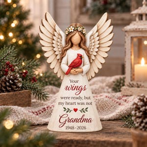 May include: A cream-colored angel figurine with large carved wings, wearing a floral crown and holding a bright red cardinal. The dress features the text: Your wings were ready, but my heart was not Grandma 1948-2026, with a small red heart.