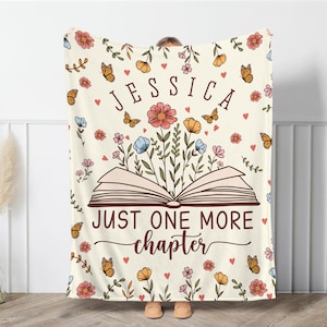 May include: Cream-colored blanket with floral and butterfly patterns. The blanket features an open book design with flowers and the text "JESSICA" and "JUST ONE MORE chapter".