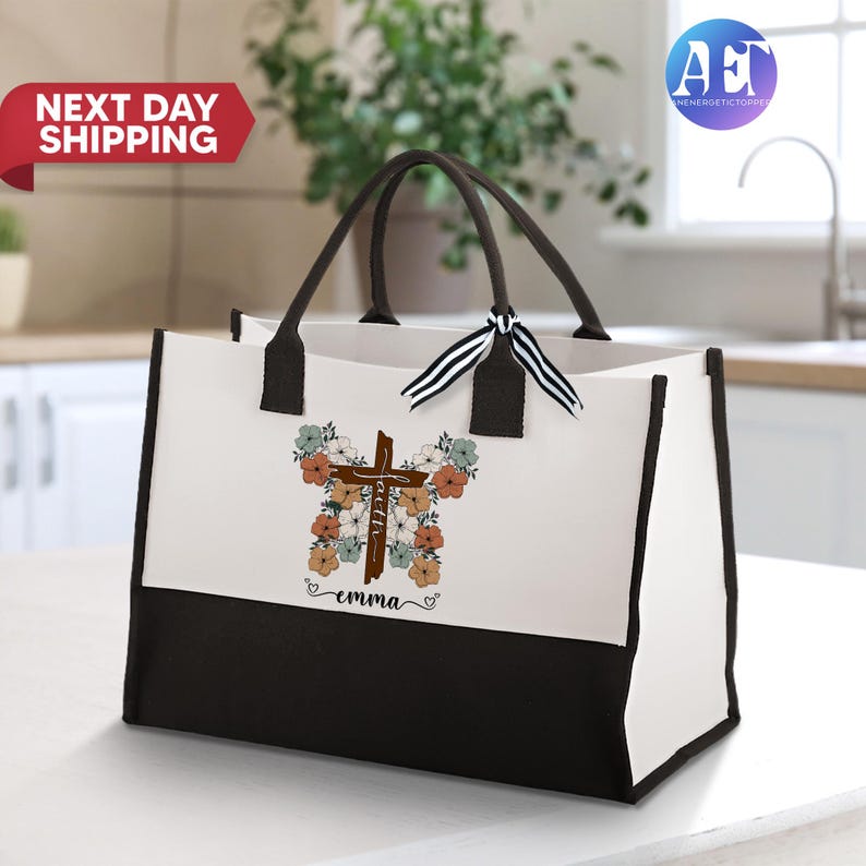 May include: A white canvas tote bag with black handles and a black bottom. The bag features a brown wooden cross with the word "faith" written on it. The cross is surrounded by colorful flowers. The name "emma" is written below the cross.