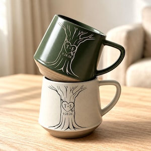 May include: Two ceramic mugs stacked vertically. The top mug is dark olive green with a white tree illustration, a heart containing the letters T+S, and the date 9.28.26. The bottom mug is off-white with a black tree illustration, heart, and date.