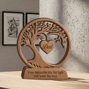 May include: A circular wooden ornament featuring a laser-cut tree design with a heart-shaped pendant hanging from the branches. The heart is engraved with the word "Dad." The base is engraved with the text "Your memories are the light that leads my way."