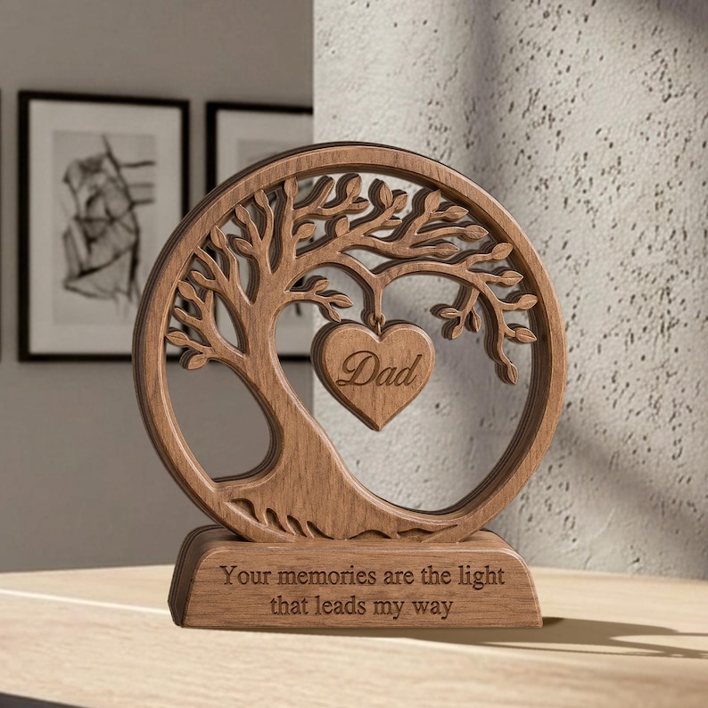 May include: A circular wooden ornament featuring a laser-cut tree design with a heart-shaped pendant hanging from the branches. The heart is engraved with the word "Dad." The base is engraved with the text "Your memories are the light that leads my way."