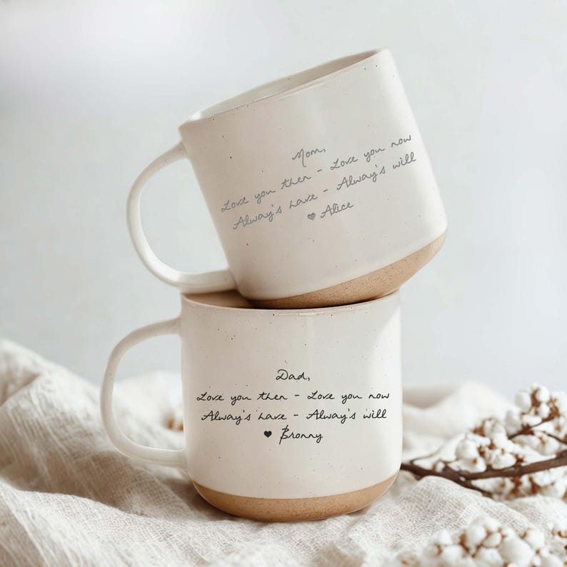 May include: Two stacked ceramic mugs with a cream-colored finish and a brown base. Each mug has handwritten text. The top mug reads "Mom" and the bottom mug reads "Dad". The mugs have a curved handle.
