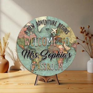 May include: A round wooden sign with a world map design and the text "A New Adventure Begins Welcome to Mrs. Sophia's Classroom." The sign is supported by a wooden stand. A small airplane graphic is also included.