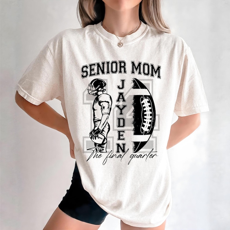May include: Cream-colored t-shirt with a black and white football-themed graphic. The design includes the text "SENIOR MOM" above a football and a football player, with the name "JAYDEN" and the phrase "The final quarter".