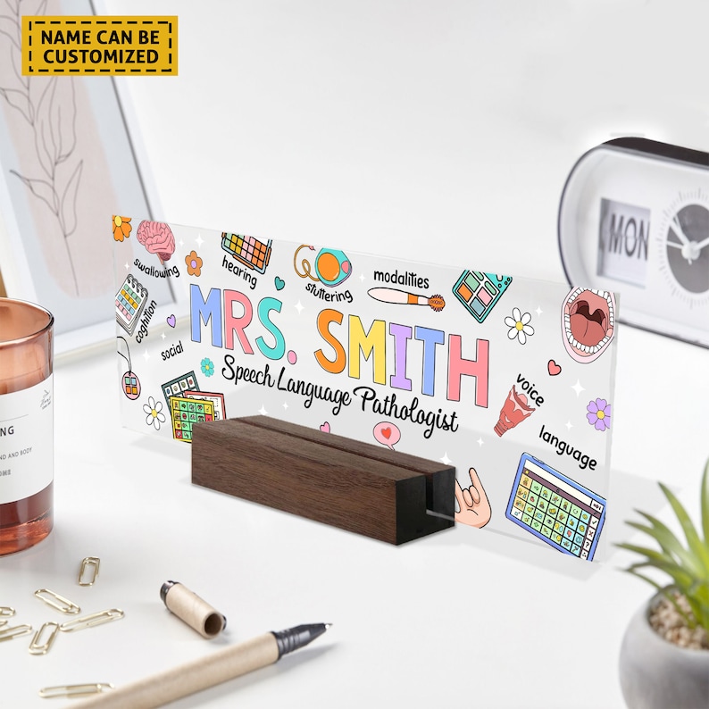 May include: A white acrylic desk sign with a colorful illustration of speech therapy symbols and the text "MRS. SMITH Speech Language Pathologist". The sign is on a wooden stand.