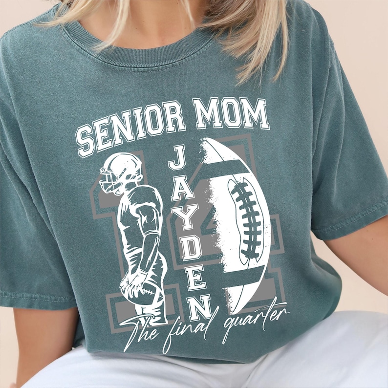 May include: Teal-colored t-shirt with the text "SENIOR MOM" in white block letters. A football graphic and the name "JAYDEN" are also featured. The phrase "The final quarter" is printed at the bottom. The shirt has a relaxed fit.