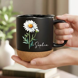 May include: Black ceramic mug featuring a daisy floral design and the name "Sophia" in white script. The mug has a cork base and a black handle. The mug is held in a person's hands.