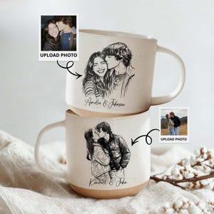 May include: Two stacked ceramic mugs with custom black and white sketch portraits of couples kissing. The mugs have a cream-colored finish with a tan base and white handle. The names "Amelia & Johnson" and "Kendall & John" are written on the mugs.