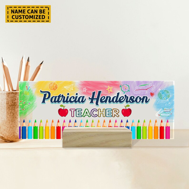 Może przedstawiać: Kolorowa szklana tabliczka na biurko z imieniem "Patricia Henderson" i napisem "TEACHER" w kolorze niebieskim. Tabliczka jest ozdobiona kolorowymi rysunkami przybor&oacute;w szkolnych, w tym oł&oacute;wk&oacute;w, globusa, książki i jabłka. Tabliczka stoi na drewnianym stojaku.