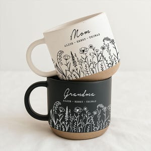May include: Two ceramic mugs stacked on top of each other. The top mug is white with the word "Mom" and floral illustrations. The bottom mug is black with the word "Grandma" and floral illustrations. Both mugs have a tan base and white floral designs.