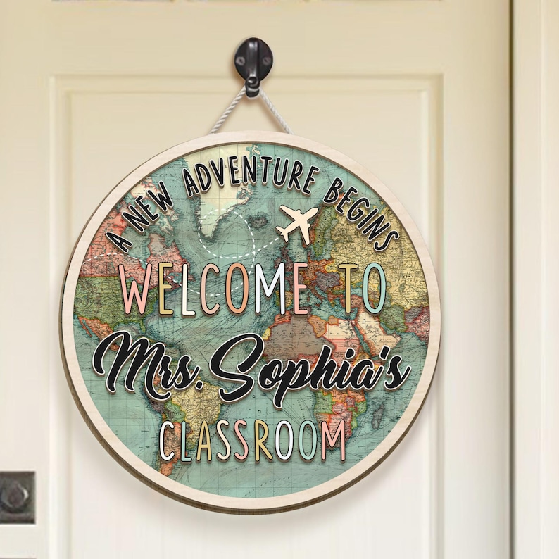 May include: Round wooden sign with a world map design. The sign reads "A NEW ADVENTURE BEGINS WELCOME TO Mrs. Sophia's CLASSROOM" in various colors. A small airplane graphic is also included.