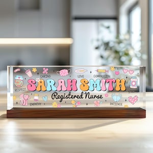 May include: A clear acrylic desk nameplate with a wood base. The nameplate features a colorful design with a nurse's uniform, medical supplies, and the words "Registered Nurse". The nameplate is personalized with the name "SARAH SMITH".
