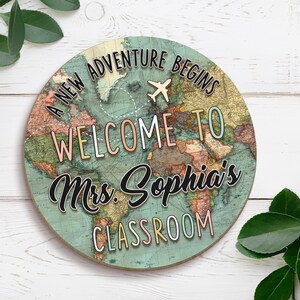 May include: Round wooden sign with a world map design. The sign reads "A NEW ADVENTURE BEGINS" and "WELCOME TO Mrs. Sophia's CLASSROOM" with a small airplane graphic. The map is in shades of blue, green, and brown.