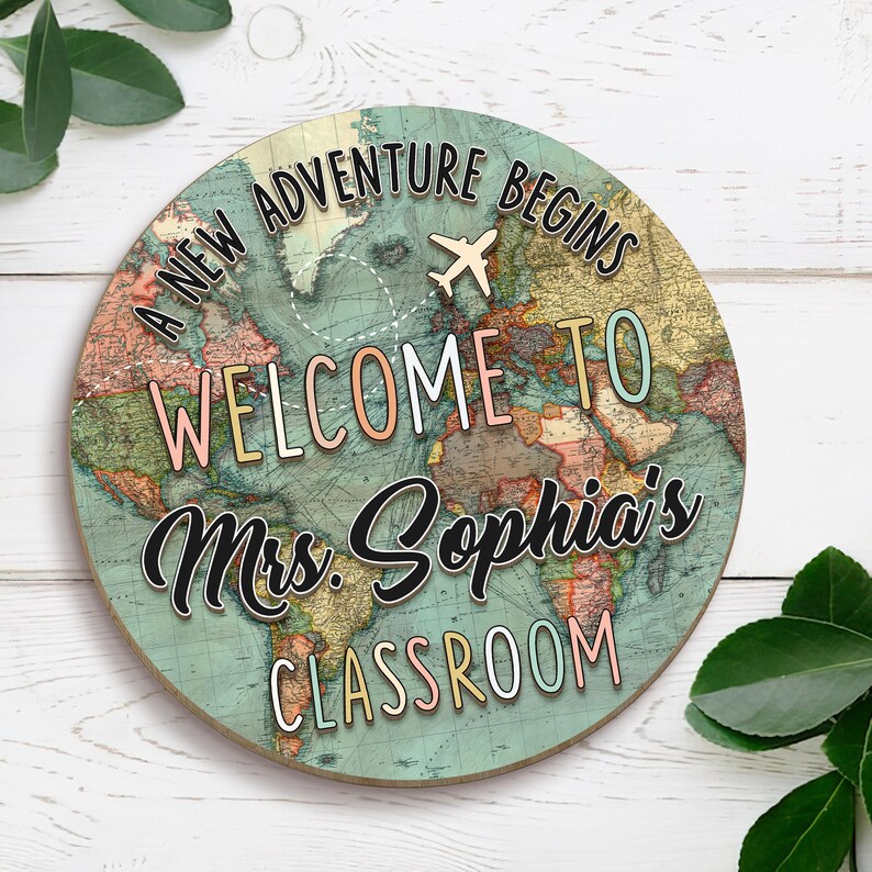 May include: Round wooden sign with a world map design. The sign reads "A NEW ADVENTURE BEGINS" and "WELCOME TO Mrs. Sophia's CLASSROOM" with a small airplane graphic. The map is in shades of blue, green, and brown.