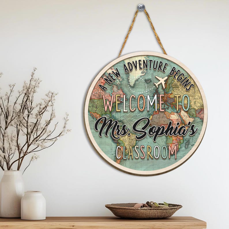 May include: Round wooden sign with a world map design. The sign reads "A New Adventure Begins, Welcome to Mrs. Sophia's Classroom" with an airplane graphic. The sign is hanging by a rope.