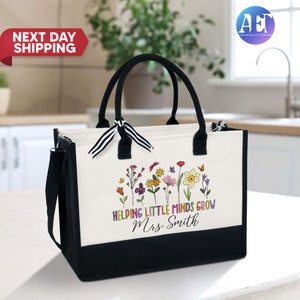Może przedstawiać: Biała torba typu tote z pł&oacute;tna z czarnymi uchwytami i wykończeniem. Torba ma kwiatowy wz&oacute;r z motylami i tekstem "Helping Little Minds Grow Mrs. Smith".