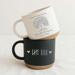 May include: Two ceramic mugs stacked on top of each other. The top mug is off-white with a willow tree design and the text "I Willow Ways Love You" and "Happy 9th Anniversary Laura & Nick". The bottom mug is black with white tally marks and hearts.