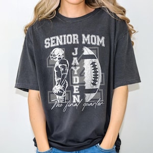 May include: Dark gray t-shirt with the text "SENIOR MOM" above a football graphic and the name "JAYDEN". A football player graphic is on the left. The text "The final quarter" is below the name. The shirt is a comfortable, casual style.
