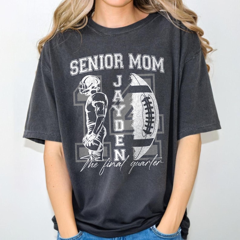 May include: Dark gray t-shirt with the text "SENIOR MOM" above a football graphic and the name "JAYDEN". A football player graphic is on the left. The text "The final quarter" is below the name. The shirt is a comfortable, casual style.