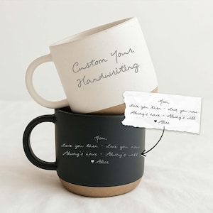 May include: Two ceramic mugs, one cream-colored and one black, stacked together. The cream mug has "Custom Your Handwriting" written on it. The black mug has a handwritten message. A note with a handwritten message is also present.