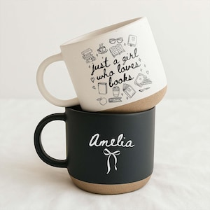 May include: Two stacked ceramic mugs. The top mug is off-white with the phrase "just a girl who loves books" and book-themed illustrations. The bottom mug is black with the name "Amelia" and a bow design.