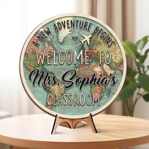 May include: A round wooden sign with a world map design. The sign reads "A NEW ADVENTURE BEGINS WELCOME TO Mrs. Sophia's CLASSROOM" with a small airplane graphic. The sign is displayed on a wooden stand.