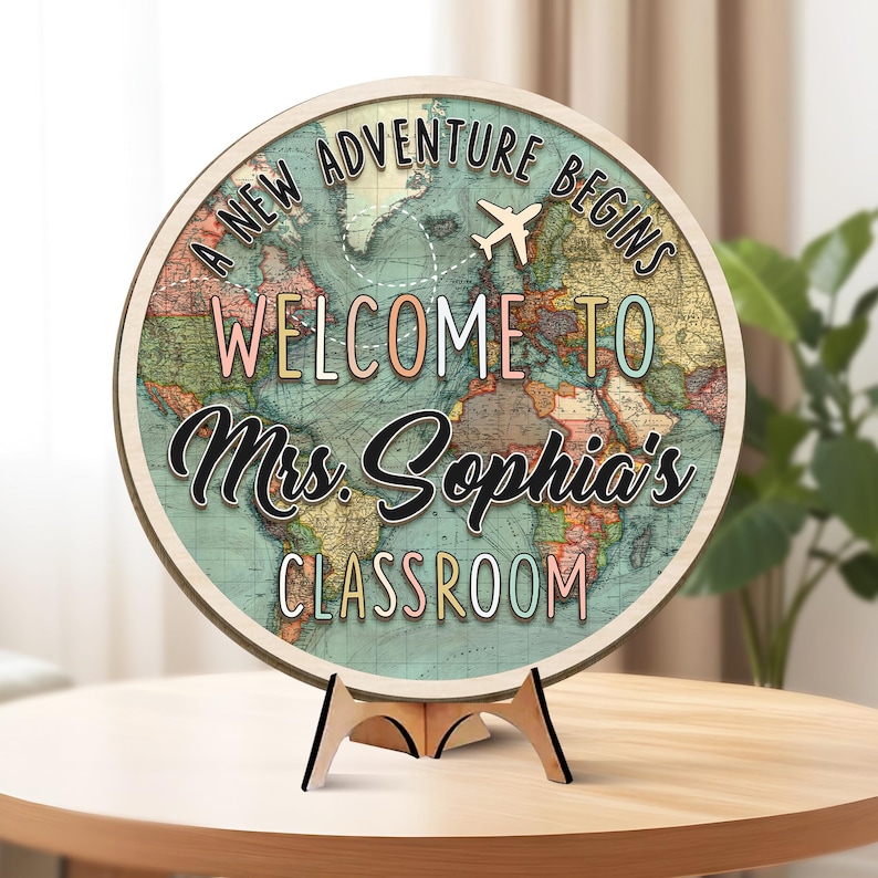 May include: A round wooden sign with a world map design. The sign reads "A NEW ADVENTURE BEGINS WELCOME TO Mrs. Sophia's CLASSROOM" with a small airplane graphic. The sign is displayed on a wooden stand.