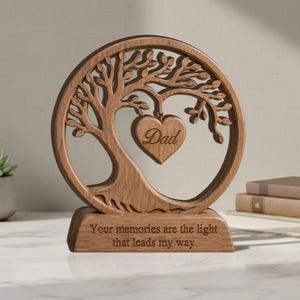 May include: A circular wooden ornament featuring a laser-cut tree design with a heart-shaped pendant in the centre engraved with the word Dad. The base is rectangular and engraved with the text Your memories are the light that leads my way.