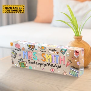 May include: A clear acrylic desk nameplate with a colorful design featuring speech therapy related icons and the text "MRS. SMITH Speech Language Pathologist".