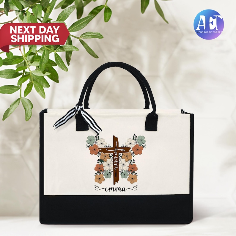 May include: A white canvas tote bag with black handles and a black bottom. The bag has a floral design with a brown cross in the center. The cross has the word "faith" written on it. The bag also has the name "emma" written below the cross.
