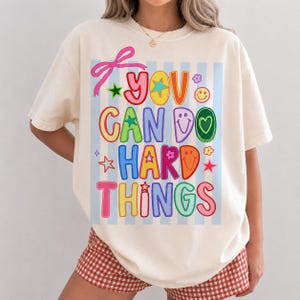 May include: A cream-colored t-shirt featuring a graphic print with light blue vertical stripes. The text reads YOU CAN DO HARD THINGS in colorful, bold letters, accented by a pink bow, stars, flowers, and smiley faces in various bright colors.