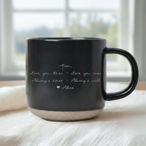 May include: Black ceramic mug with a rounded base and a large handle. The mug features handwritten text that reads "Mom, Love you then - Love you now, Always have - Always will, Alice". The base is a light tan color.