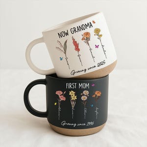 May include: Two ceramic mugs, one white and one black, stacked. The white mug reads "NOW GRANDMA" and features floral illustrations and names. The black mug says "FIRST MOM" with similar floral designs and names. Both mugs have a brown base and white handles.
