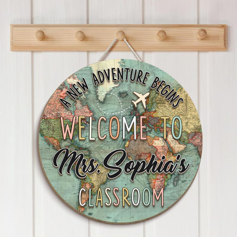 May include: A round wooden sign with a world map design and the text "A New Adventure Begins Welcome to Mrs. Sophia's Classroom". The sign has a white rope for hanging and is mounted on a wooden rack with pegs.