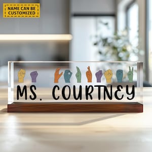 May include: A clear acrylic desk nameplate with a wooden base. The nameplate features a row of colorful hand signs in American Sign Language and the text "MS. COURTNEY".