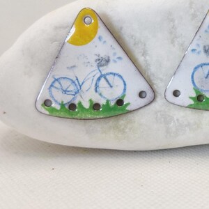 May include: A pair of white triangle-shaped earrings with a blue bicycle design, a yellow sun, and green grass. The earrings have a hole at the top for hanging.