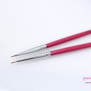 May include: Two fine-tipped paint brushes with magenta handles and silver ferrules. The brushes are angled slightly, showcasing their fine tips. The brand name "pearly karpel" is printed on the handles. The image is on a white background, highlighting the brushes.