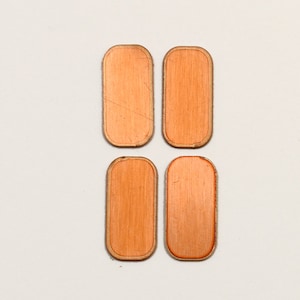 May include: Four copper-colored, rectangular coasters with rounded corners are arranged in a square. Each coaster has a visible wood grain pattern and a thin, darker border. The coasters are set against a plain white background.
