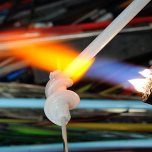 May include: A white glass rod is being heated with a torch. The torch has a blue flame and is being held by a person's hand, which is not visible in the image. The glass rod is being heated to a molten state, and the flame is creating a spiral shape in the glass.