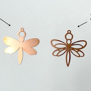 May include: Two copper-colored dragonfly pendants. One is solid, the other is an openwork design. Both have a small loop at the top for hanging. The dragonflies are displayed against a white background.